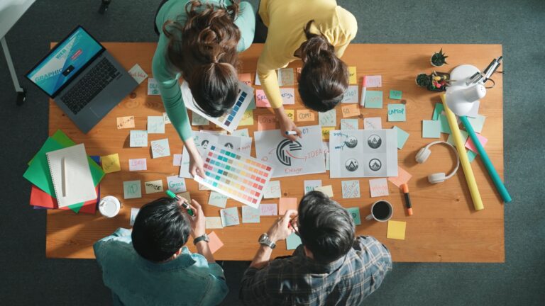 Vier Personen arbeiten von oben gesehen an einem Tisch voller Haftnotizen an Logo-Entwürfen, mit Farbfächern und Skizzen