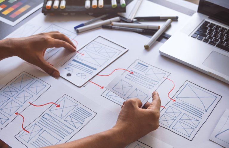 Hände ordnen skizzierte Wireframes einer App auf einem Tisch, daneben Laptop und Stifte